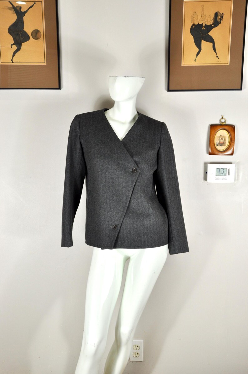 May include: A dark gray blazer with a V-neck and long sleeves. The blazer has a wrap-style front with two buttons. The fabric has a subtle herringbone pattern. The blazer is displayed on a white mannequin.