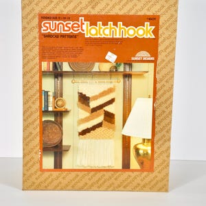 May include: A Sunset Latch Hook kit box titled Shadow Patterns featuring a finished wall hanging with geometric brown, tan, and cream yarn segments and long fringe. The box displays a finished size of 15 inches by 39.5 inches on the front panel.