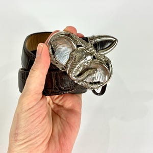 May include: A brown leather belt with a silver elephant-shaped buckle. The buckle is detailed with the elephant's head and tusks. The belt is held in a hand against a white background.
