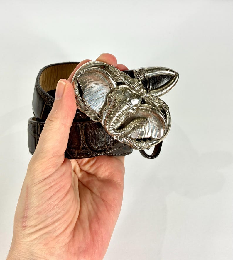 May include: A brown leather belt with a silver elephant-shaped buckle. The buckle is detailed with the elephant's head and tusks. The belt is held in a hand against a white background.