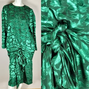 Op de afbeelding: Smaragdgroene jurk met lange mouwen en een bloemenpatroon. De jurk heeft een ronde hals en een knoopdetail in de taille. De stof heeft een subtiele glans en de jurk wordt op een mannequin gepresenteerd.