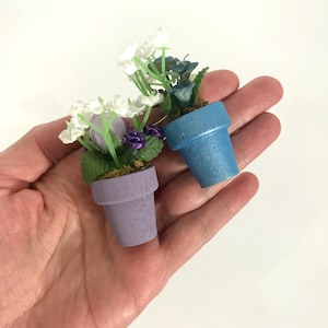 Pode incluir: Duas plantas em vaso miniatura com flores brancas e roxas. Os vasos são pintados de roxo claro e azul claro.