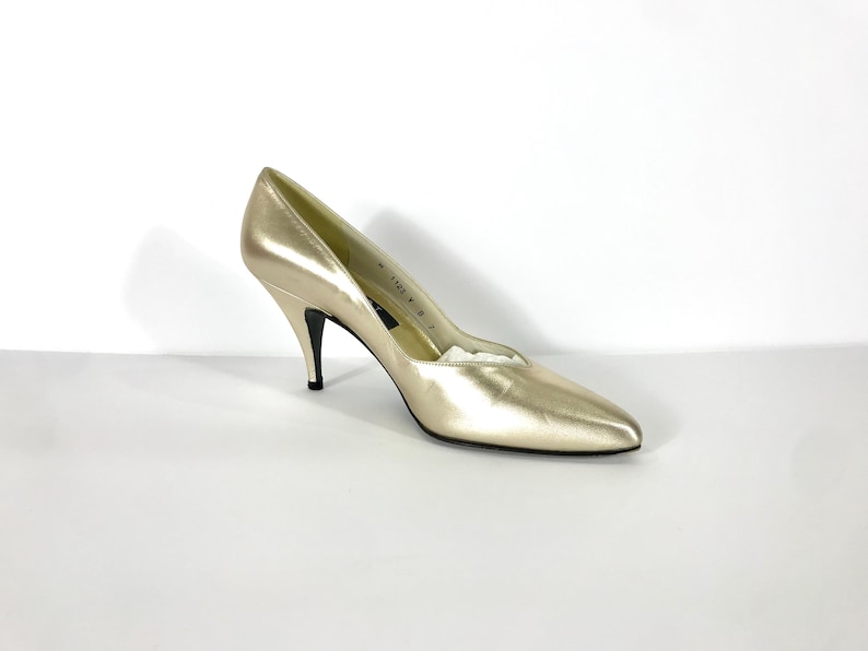 May include: A single gold-colored high heel shoe with a pointed toe and a thin, black heel. The shoe is made of a shiny material and is set against a white background. The heel measures approximately 3 inches.