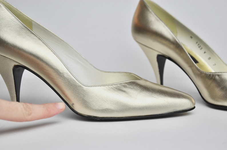 May include: A pair of gold-colored high heels with pointed toes and stiletto heels. The shoes have a metallic sheen and a classic design, suitable for formal occasions. The heels are approximately 3 inches tall.