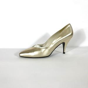 May include: A single gold-colored high heel shoe with a pointed toe and a stiletto heel. The shoe is made of a shiny material and is set against a white background. The heel is approximately 3 inches high.