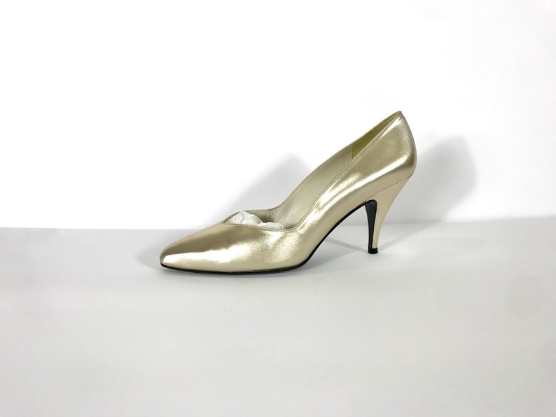 May include: A single gold-colored high heel shoe with a pointed toe and a stiletto heel. The shoe is made of a shiny material and is set against a white background. The heel is approximately 3 inches high.