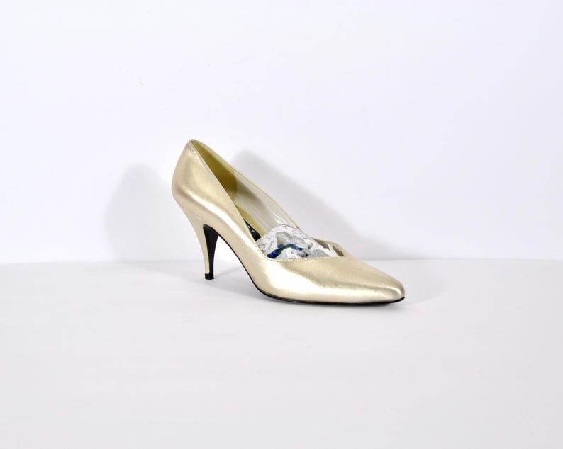 May include: A single gold-colored high heel shoe with a pointed toe and a thin black heel. The shoe is made of a shiny material and is set against a white background.