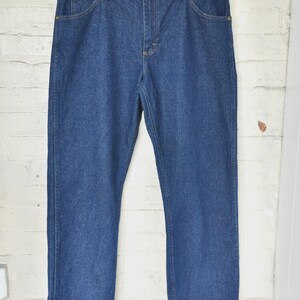 May include: A pair of blue denim jeans hanging on a black hanger. The jeans have a classic five-pocket design with a button and zip closure. The denim is a medium wash, and the jeans appear to be new.