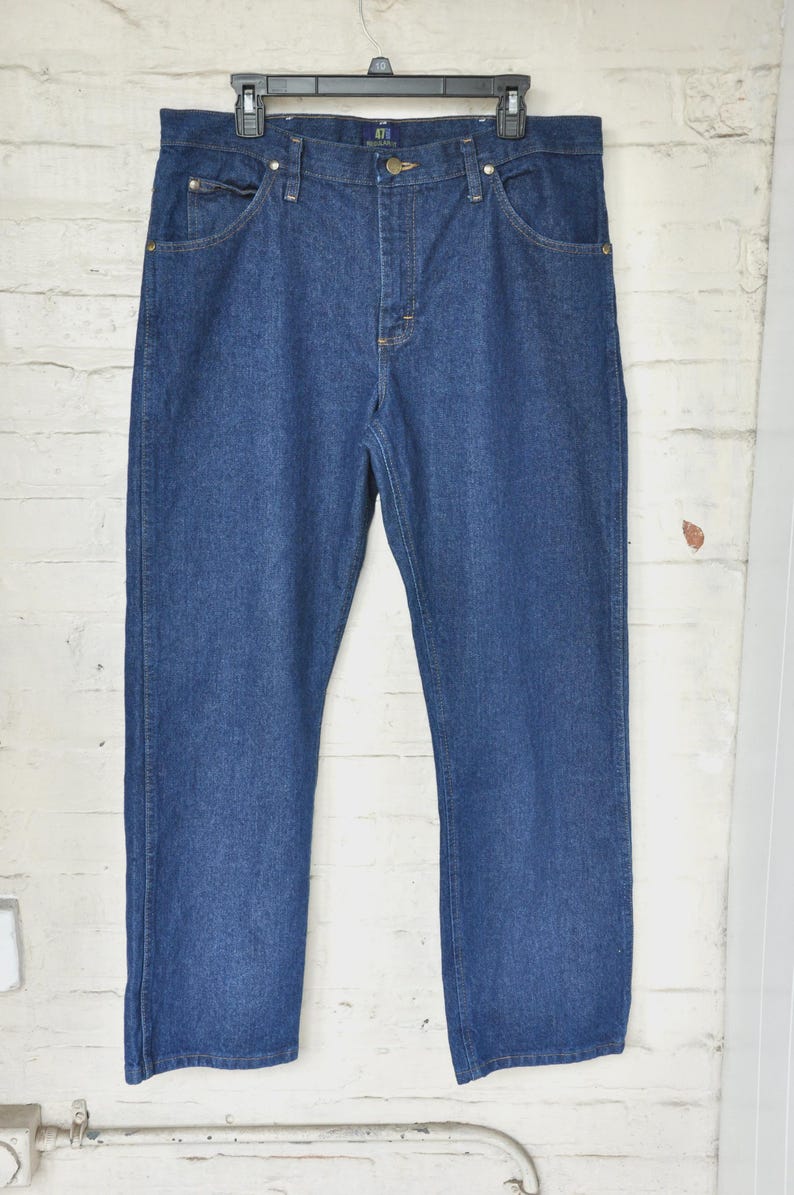 May include: A pair of blue denim jeans hanging on a black hanger. The jeans have a classic five-pocket design with a button and zip closure. The denim is a medium wash, and the jeans appear to be new.