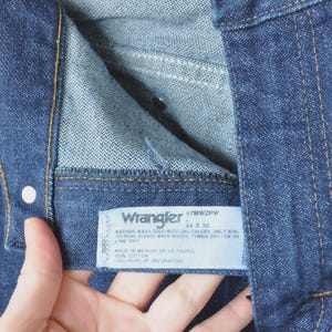 May include: Close-up of blue denim jeans with a visible label. The label reads "Wrangler" and includes size information, 34 x 30. The jeans have a button and stitching details.