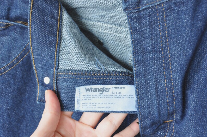 May include: Close-up of blue denim jeans with a visible label. The label reads "Wrangler" and includes size information, 34 x 30. The jeans have a button and stitching details.