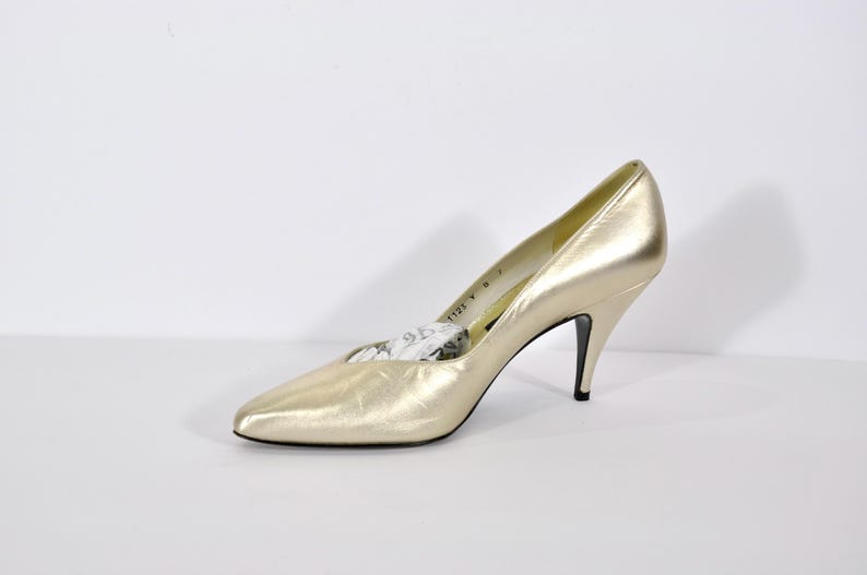 May include: A single gold-colored high heel shoe with a pointed toe and a thin black heel. The shoe has a metallic sheen and appears to be made of leather. The heel is approximately 3 inches tall.