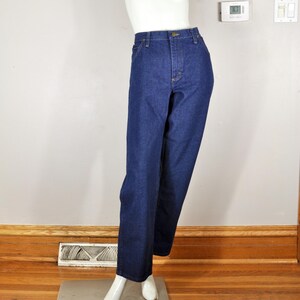 May include: Dark blue denim jeans on a mannequin. The jeans have a straight-leg cut, a button and zip fastening, and pockets. The mannequin is white and stands on a metal plate, against a neutral wall and wooden floor.