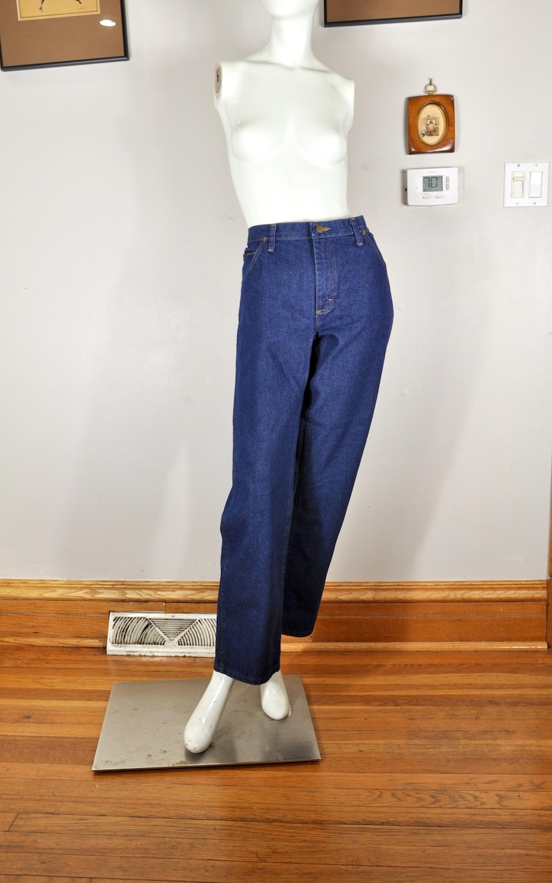 May include: Dark blue denim jeans on a mannequin. The jeans have a straight-leg cut, a button and zip fastening, and pockets. The mannequin is white and stands on a metal plate, against a neutral wall and wooden floor.