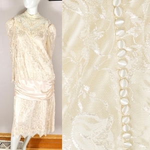 Puede incluir: Vestido de encaje marfil con cuello alto y mangas largas. El vestido presenta una cinturilla de satén y una fila de botones cubiertos de tela en la parte delantera. El vestido se exhibe en un maniquí.