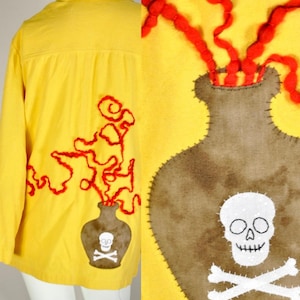 Può includere: Una camicia gialla brillante con bottoni e una grande applicazione a forma di bottiglia marrone sul retro. La bottiglia presenta un teschio bianco con ossa incrociate e decorazioni in filo rosso che fuoriescono, cucite con filo scuro visibile.