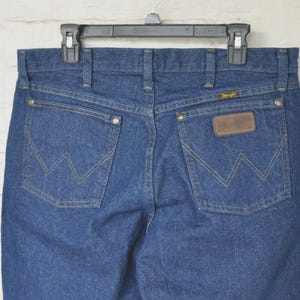 May include: Blue denim jeans with the Wrangler logo on the back pockets and a brown leather patch. The jeans are hanging on a black hanger against a white brick wall.