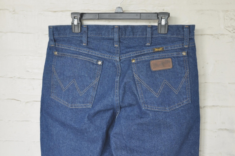 May include: Blue denim jeans with the Wrangler logo on the back pockets and a brown leather patch. The jeans are hanging on a black hanger against a white brick wall.