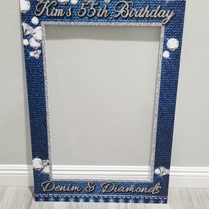 May include: A blue and silver photo booth prop with a denim and diamond theme. The text "Kim's 55th Birthday" and "Denim & Diamonds" are printed on the prop.