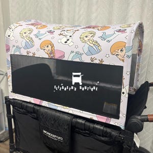 May include: A white and light pink canopy with a black mesh window, decorated with cartoon characters and stars. The canopy is attached to a black Wonderfold wagon. The text "Elizabeth's Boutique" is visible on the window.