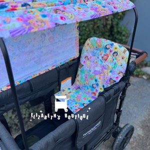 May include: A black wagon with a canopy and two seats. The canopy is made of a white fabric with a colorful floral print featuring princesses. The wagon has a Wonderfold Wagon logo on the side. The wagon is on a paved surface.