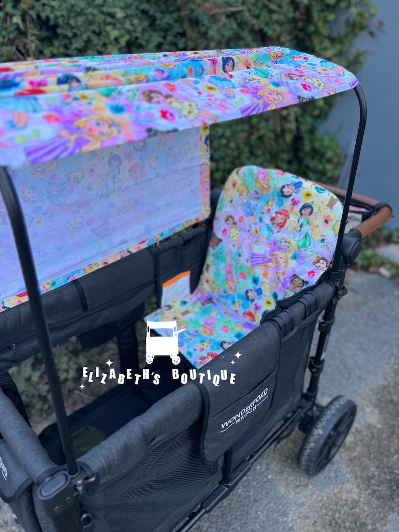 May include: A black wagon with a canopy and two seats. The canopy is made of a white fabric with a colorful floral print featuring princesses. The wagon has a Wonderfold Wagon logo on the side. The wagon is on a paved surface.