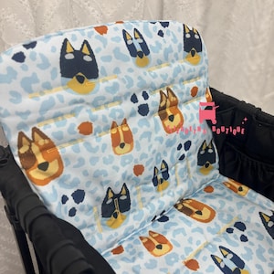 May include: A blue and white stroller seat liner with a pattern of brown and black dog faces. The liner has a padded back and seat.