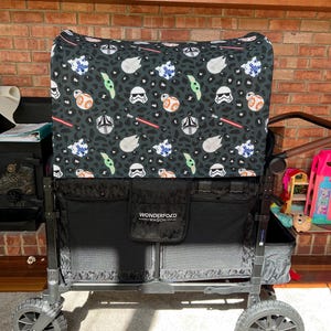 May include: A black and grey WonderFold wagon with a patterned canopy featuring Star Wars characters. The canopy has images of Stormtroopers, BB-8, and lightsabers. The wagon has large grey wheels and a black storage compartment.