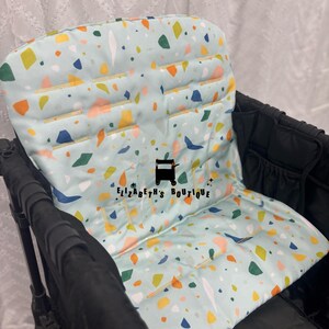 May include: A light blue stroller seat liner with a terrazzo pattern. The liner has a padded back and seat area. The liner is made of fabric and has a zipper closure. The liner is for a stroller and is designed to protect the stroller seat from dirt and spills. The liner is also comfortable for the child to sit on. The liner has the text "Elizabeth's Boutique" printed on it.