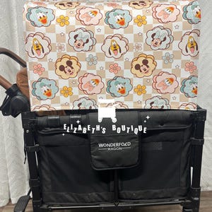 May include: A black wagon with a white and brown fabric canopy featuring a checkered pattern with cartoon images of Mickey Mouse, Minnie Mouse, and Donald Duck. The canopy has the text "WONDERFOLD WAGON" on the front. The wagon is on a wooden floor.