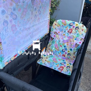 May include: A black wagon with a canopy and a seat. The canopy and seat are covered in a white fabric with a colorful print of Disney princesses. The wagon has a black mesh storage area and the text "WONDER" is visible on the side.