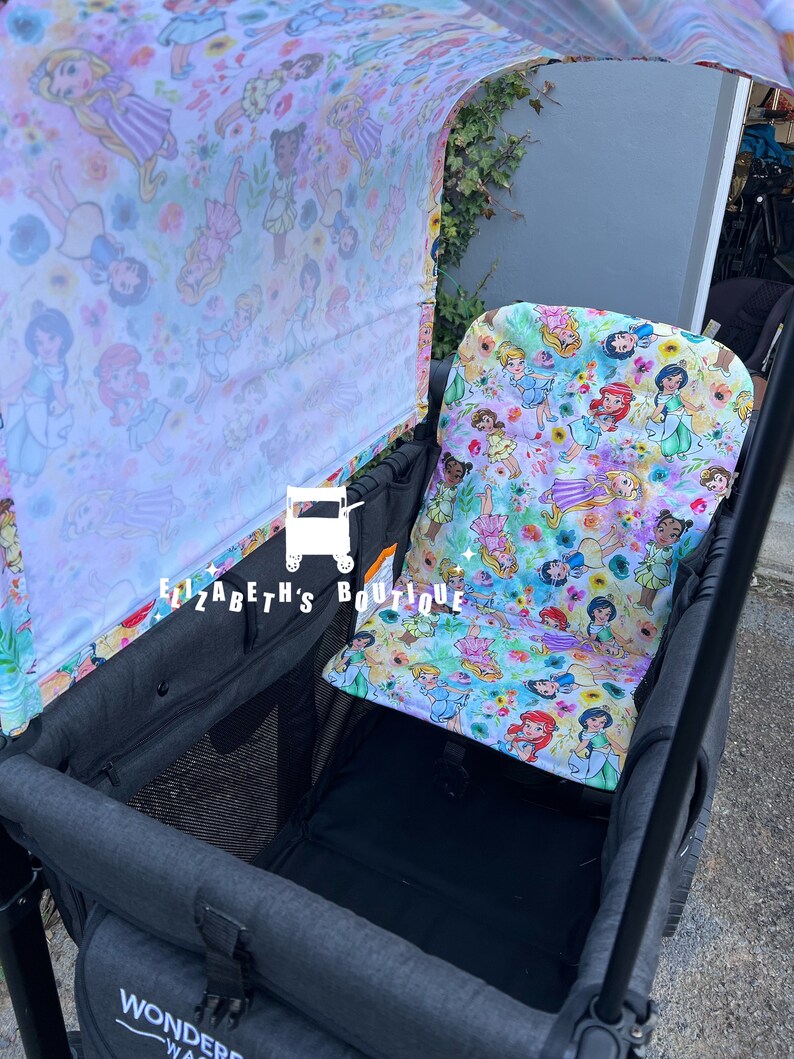May include: A black wagon with a canopy and a seat. The canopy and seat are covered in a white fabric with a colorful print of Disney princesses. The wagon has a black mesh storage area and the text "WONDER" is visible on the side.