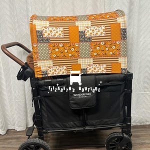 May include: A black Wonderfold wagon with a patchwork fabric cover in shades of orange, brown, and white. The cover features a floral pattern and is designed to fit over the wagon's back.