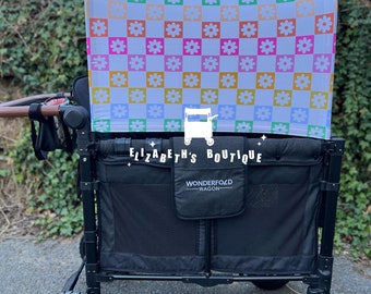 May include: A black wagon with a white and colourful checkered canopy featuring a floral pattern. The canopy has the text "Elizabeth's Boutique" on it. The wagon has the text "Wonderfold Wagon" on it.