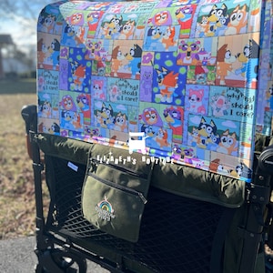 Op de afbeelding: Een groene wagen met een blauw en wit zonnedak met een cartoonhondpersonage. Het zonnedak heeft een herhalend patroon van het hondpersonage met de tekst "Why should I care?" De wagen heeft een groene zak met de tekst "Rainbow Baby" erop.