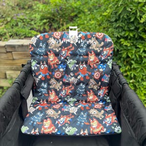 May include: Black stroller seat cover with a colorful pattern of cartoon characters in superhero costumes. The cover is inside a black stroller, with a white plastic clip at the top. The background is green foliage.