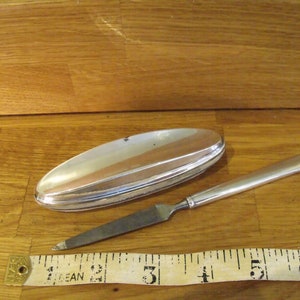 May include: A silver letter opener and a matching oval-shaped container. The letter opener has a long, thin blade and a handle. The container is closed and has a curved top. A measuring tape is in the foreground.