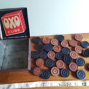 May include: A vintage OXO cube tin, featuring a red and white logo, sits open. Scattered around are wooden draughts pieces in brown and black. The tin's lid displays the text "OXO CUBE" and "OXO STRENGTHENS EVERY DISH."