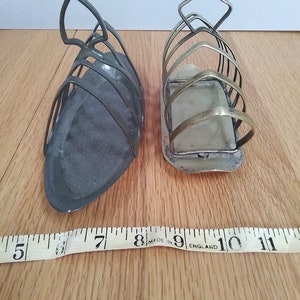 May include: Two vintage metal toast racks. One is dark grey, the other is silver. Each rack has a series of curved bars to hold slices of toast upright. A measuring tape is in the foreground. The racks are on a wooden surface.
