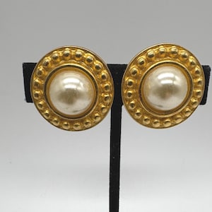May include: A pair of round, gold-toned clip-on earrings. Each earring features a large, off-white pearl center surrounded by a textured gold border with small, raised dots. The earrings are displayed on a black stand against a white background.