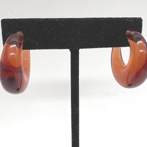 May include: A pair of hoop earrings crafted from translucent amber-colored resin. The earrings feature a thick, rounded, C-shaped design with a smooth, glossy finish that allows light to pass through the material, highlighting deep orange tones.