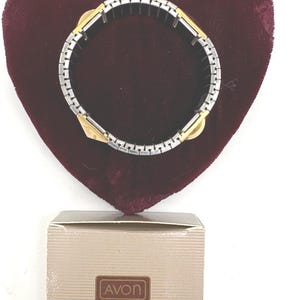 May include: A silver and gold-toned bracelet with a rectangular link design. The bracelet is displayed on a burgundy heart-shaped velvet surface. A beige box with the word "AVON" is at the bottom.