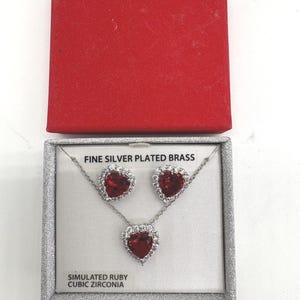 May include: A fine silver-plated brass jewelry set featuring a necklace and matching earrings. Each piece has a heart-shaped simulated ruby center stone surrounded by cubic zirconia. The set is displayed in a silver-trimmed box with a red lid.