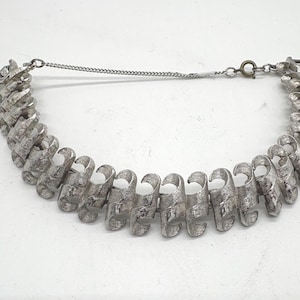 May include: Silver-toned necklace with a unique, textured design. The necklace features a series of curved, scroll-like links connected by a chain. The metal has a slightly aged appearance, adding to its vintage appeal. The necklace is a statement piece.