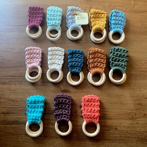 May include: A collection of 12 crocheted teethers in various colors, including teal, pink, purple, white, brown, orange, yellow, and blue. Each teether has a wooden ring attached to the crocheted fabric.