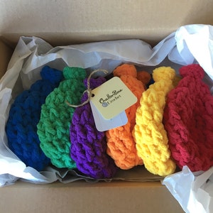 May include: A box filled with seven colorful, crocheted items arranged in a rainbow spectrum. The colors include blue, green, purple, orange, yellow, and red. A tag with the text "ChatterBox Crochet" is attached.
