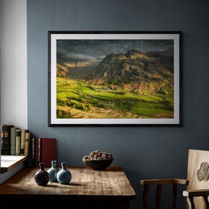 May include: Framed landscape print featuring a vibrant green valley nestled between towering mountains under a dramatic, cloudy sky. The print is displayed on a dark blue wall above a wooden table with decorative vases and a bowl of potpourri.