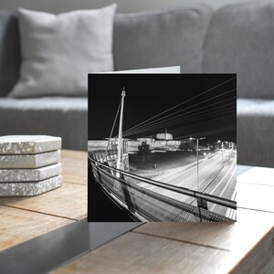 Puede incluir: Fotografía en blanco y negro de un puente moderno por la noche. La imagen muestra la arquitectura del puente, con rastros de luces de automóviles de larga exposición en la carretera de abajo. Una pila de cuatro posavasos grises y blancos descansa sobre una mesa de madera.