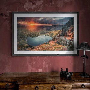 May include: A framed landscape print depicts a mountain range, lake, and sunset. The artwork is in a dark wooden frame, displayed on a wooden dresser. A black lamp and decorative ducks are on the dresser. The wall is painted a deep red.
