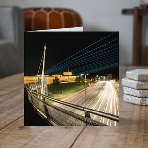 Puede incluir: Tarjeta de felicitación cuadrada con una escena nocturna de un puente y luces de la ciudad. La imagen muestra un puente con una pasarela curva y un edificio al fondo. La larga exposición captura rastros de luz de vehículos.
